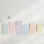 Flat-lay: a tiny potted seedling in a brass pot beside a neat row of small pastel cards standing upright like a miniature skyline on a pale off-white background