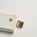 Flat-lay: a neat stack of pale paper lecture notes beside a small brass abacus with a single gold coin placed on top, on a pale off-white background