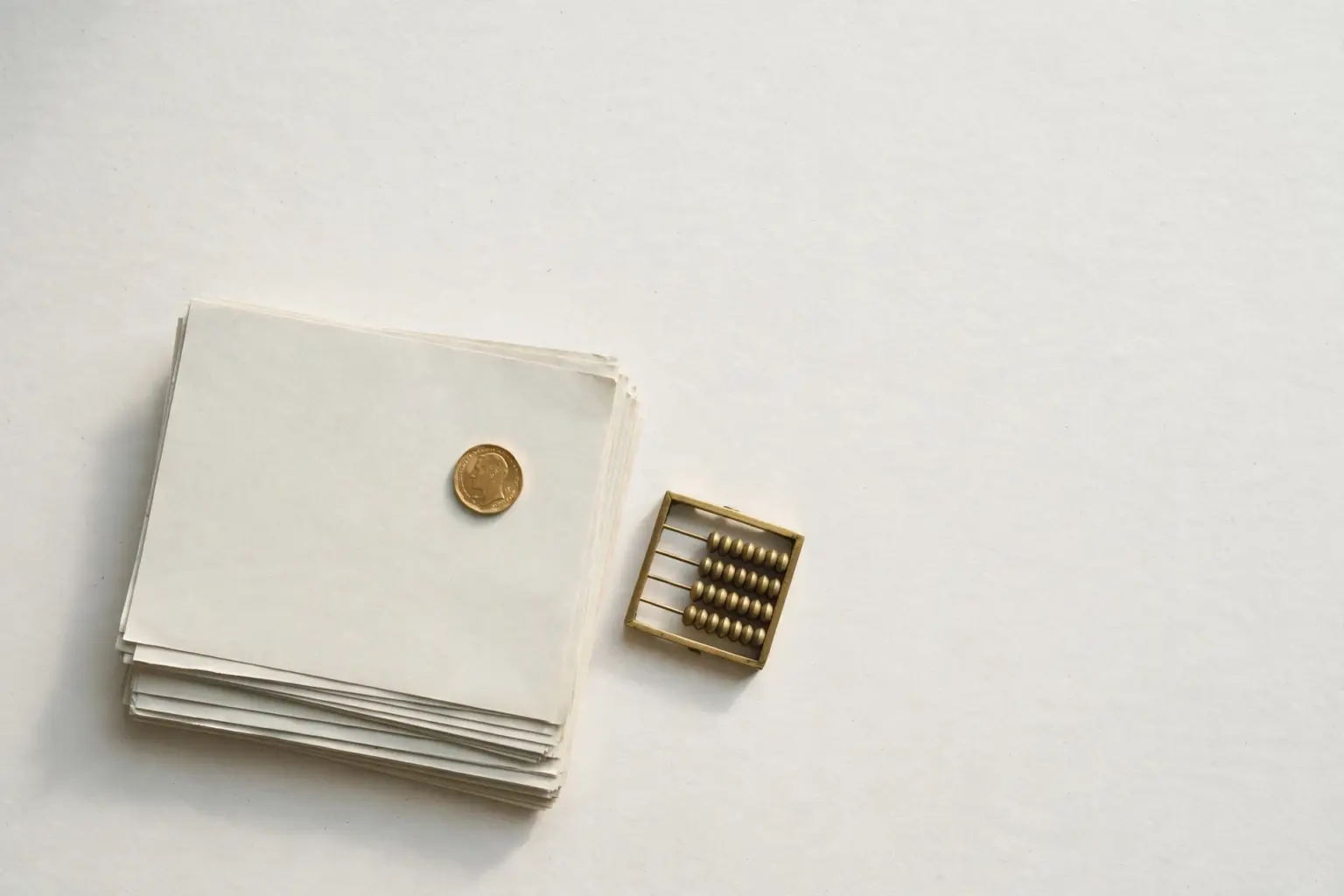Flat-lay: a neat stack of pale paper lecture notes beside a small brass abacus with a single gold coin placed on top, on a pale off-white background