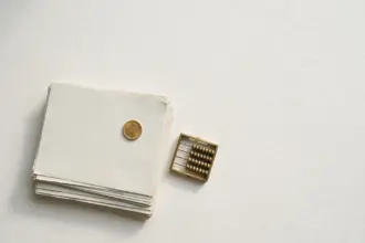 Flat-lay: a neat stack of pale paper lecture notes beside a small brass abacus with a single gold coin placed on top, on a pale off-white background